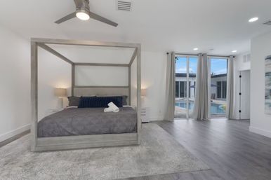 Bright modern master bedroom with gray four‑poster bed, plush rug, bedside lamps and ceiling fan, plus sliding glass doors opening to a poolside patio.