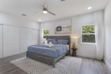 Bright modern coastal-style bedroom with a gray wood bed dressed in blue linens, patterned rug, ceiling fan, bedside lamps, white sliding closet doors and two windows revealing lush greenery.