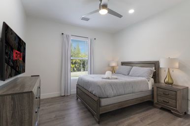 Sunlit modern bedroom with gray bedding on a wooden bed, matching nightstands and dresser, ceiling fan, wall-mounted TV showing Netflix, and a window with white curtains overlooking trees.