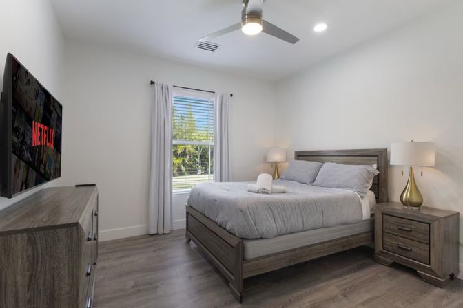 Sunlit modern bedroom with gray bedding on a wooden bed, matching nightstands and dresser, ceiling fan, wall-mounted TV showing Netflix, and a window with white curtains overlooking trees.