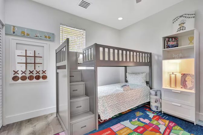 Bright children’s bedroom interior with gray bunk bed and storage stairs, patterned bedding, playful U.S. map rug, white shelving with lamp, tic‑tac‑toe wall art and window blinds.