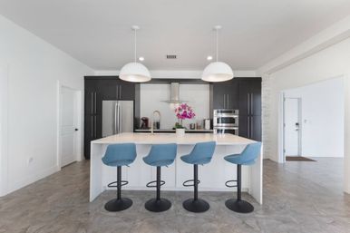 Contemporary open-plan kitchen with white island and quartz countertop, four blue swivel bar stools, dark cabinets, stainless steel fridge and double ovens, two white pendant lights and pink orchid centerpiece.