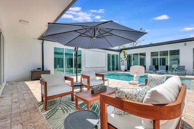 Modern backyard patio and pool deck with cushioned wooden lounge chairs, coffee table and large navy umbrella on stone pavers, rectangular swimming pool surrounded by sliding glass doors under a bright blue sky.