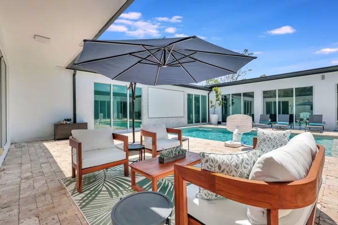Modern backyard patio and pool deck with cushioned wooden lounge chairs, coffee table and large navy umbrella on stone pavers, rectangular swimming pool surrounded by sliding glass doors under a bright blue sky.