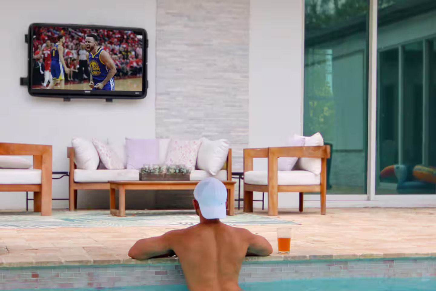 Relaxed man in a white cap leans on the edge of a backyard pool watching a wall-mounted TV broadcasting a basketball game, with wooden lounge chairs, soft cushions and a cold drink on the pool ledge.