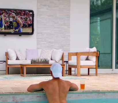 Relaxed man in a white cap leans on the edge of a backyard pool watching a wall-mounted TV broadcasting a basketball game, with wooden lounge chairs, soft cushions and a cold drink on the pool ledge.