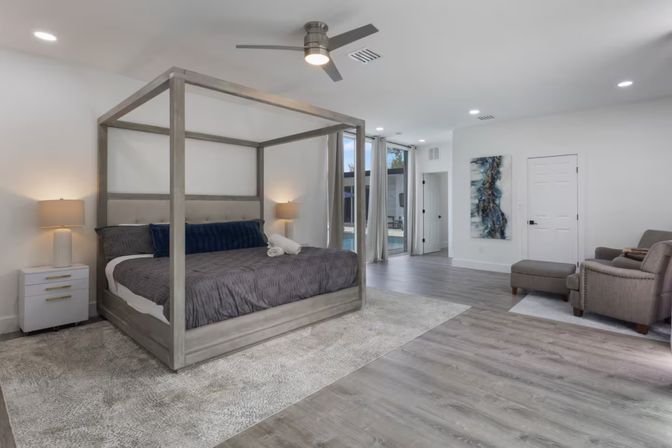 Spacious modern master bedroom with gray four-poster bed, plush rug, wood-look floors, seating area, ceiling fan and sliding glass doors to a pool patio.