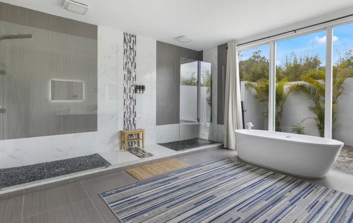 Spa-like modern bathroom with a freestanding white tub by floor-to-ceiling glass doors overlooking a palm-lined patio, dual walk-in showers with mosaic tile accents, gray floor tiles and a blue-striped rug.