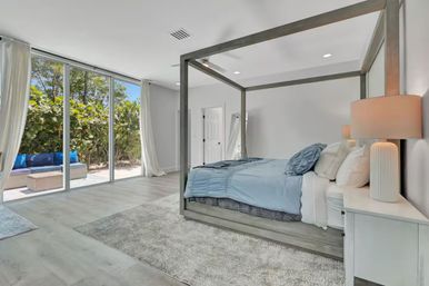 Bright, airy modern master bedroom with gray four‑poster bed and light blue bedding, plush area rug, bedside lamp on white nightstand, and floor-to-ceiling sliding glass doors opening to a sunny patio with outdoor seating and lush greenery.