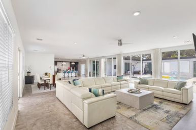 Sunlit modern open-plan living room with white leather sectional sofas and a central coffee table on a patterned rug, adjacent dining area and kitchen island with blue bar stools, floor-to-ceiling windows and sliding doors opening to a poolside patio.