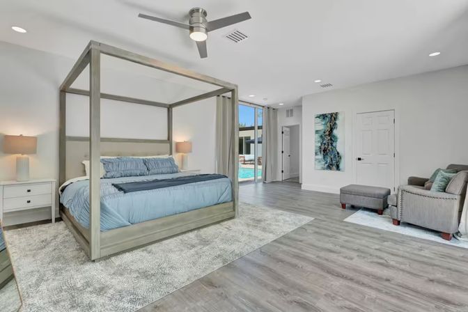 Bright, airy modern master bedroom with light-wood four-poster bed, blue bedding, matching nightstands and lamps, seating area with ottoman, wide wood-look floors and sliding glass doors revealing a backyard pool.