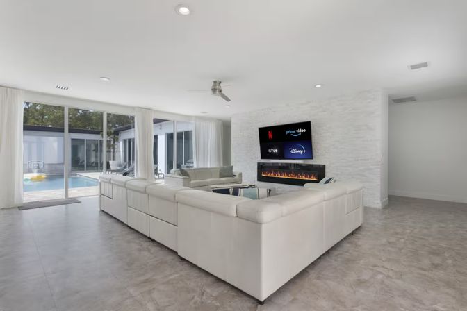 Bright modern living room with a large white leather sectional, wall-mounted flat-screen showing streaming apps above a linear electric fireplace, and floor-to-ceiling sliding glass doors opening to a sunny backyard pool.