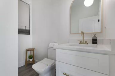 Bright modern white powder room with a white vanity, arched gold-framed mirror, brass faucet, toilet, wooden stool with potted plant, black-and-white wall art, and gray flooring.
