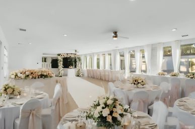 Elegant indoor wedding venue: bright banquet hall with round tables draped in white, blush-sashed chairs, rose and cream centerpieces, candles, a white aisle leading to a floral arch, and large sunlit windows