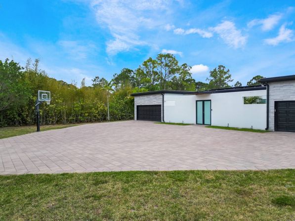 Modern single-story home with double black garage doors, expansive paved driveway and freestanding basketball hoop, grassy lawn and tree-lined yard under a bright blue sky.