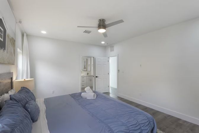Bright airy modern master bedroom with blue bedding, ceiling fan, hardwood floors and open en-suite bathroom; rolled towels on the bed, white walls and bedside lamp.