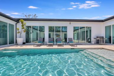 Modern courtyard with turquoise swimming pool, four lounge chairs with teal cushions, sliding glass doors, potted plant and stainless gas grill under a bright blue sky