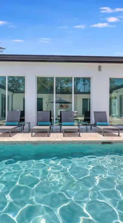 Modern courtyard with turquoise swimming pool, four lounge chairs with teal cushions, sliding glass doors, potted plant and stainless gas grill under a bright blue sky