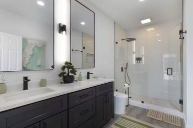 Bright contemporary bathroom with black double-sink vanity and white quartz countertop, two tall mirrors and black faucets, potted bonsai, frameless glass walk-in shower with rain and handheld fixtures, light wood-look flooring and green striped bath rug.
