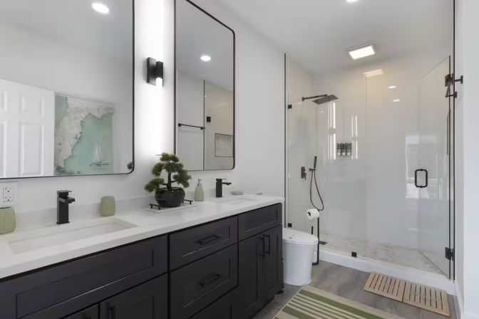 Bright contemporary bathroom with black double-sink vanity and white quartz countertop, two tall mirrors and black faucets, potted bonsai, frameless glass walk-in shower with rain and handheld fixtures, light wood-look flooring and green striped bath rug.