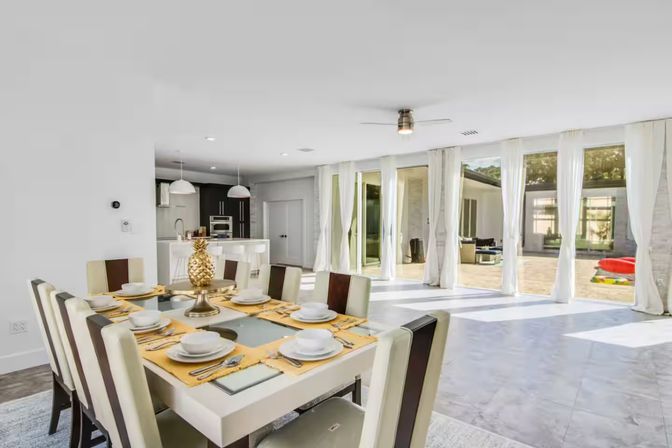 Bright modern open-plan dining room and kitchen with large sliding glass doors to a sunlit patio, white sheer curtains, tile floor, set table with yellow placemats and gold pineapple centerpiece