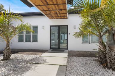 Sunny modern white coastal home entrance with glass double doors, wood-paneled overhang, concrete paver walkway, palm trees and white gravel landscaping.