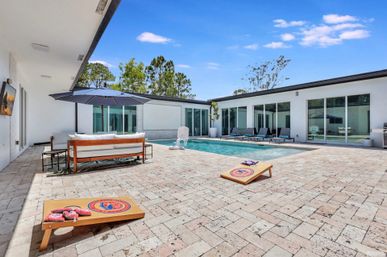 Modern private backyard patio with rectangular swimming pool, sun loungers and umbrella-shaded sofa, cornhole boards on a stone paver deck and floor-to-ceiling sliding glass doors under a clear blue sky.
