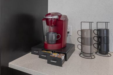 Cozy countertop coffee station: red single-serve coffee maker on speckled kitchen counter with open drawer of K-cups and two vertical mug racks holding neutral-tone mugs