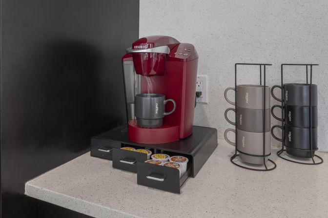 Cozy countertop coffee station: red single-serve coffee maker on speckled kitchen counter with open drawer of K-cups and two vertical mug racks holding neutral-tone mugs