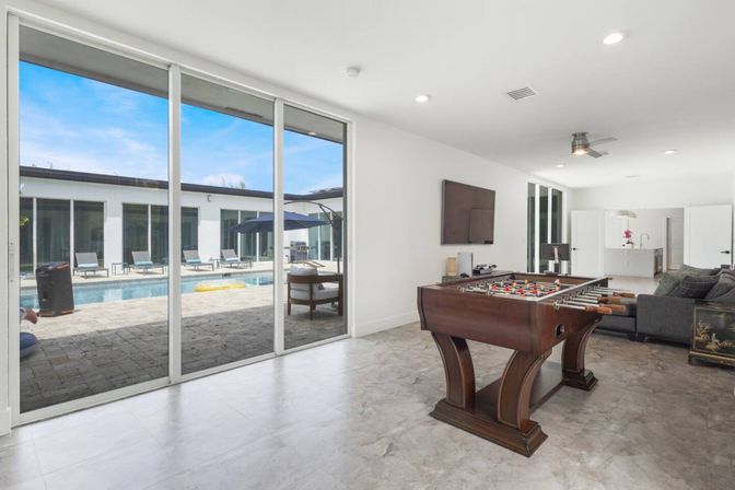 Modern game room with a wooden foosball table and large sliding glass doors opening to a sunny poolside patio with lounge chairs and umbrella.