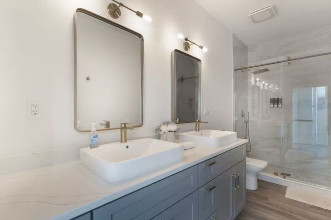 Bright modern bathroom with double white vessel sinks on a marble-look countertop, gray shaker vanity, brass faucets and rectangular mirrors with wall sconces, glass-enclosed walk-in shower and wood-look flooring — sleek, spa-like design.