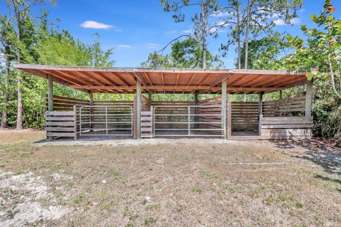 Rustic open-air wooden three-stall shelter with metal gates on a grassy lot, surrounded by trees under a blue sky.