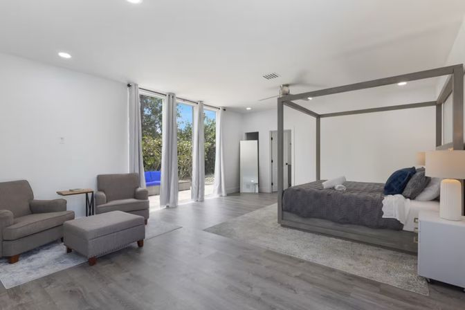 Sunlit modern master suite with gray four-poster bed, cozy sitting area with armchairs and ottoman, floor-to-ceiling windows opening to patio, light wood floors.