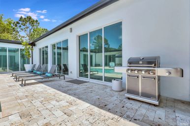 Sunny modern backyard pool patio with travertine pavers, large sliding glass doors, four gray chaise lounges with turquoise neck rolls, stainless-steel gas grill and pool reflecting trees under a bright blue sky.