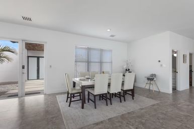 Bright modern dining room with white walls, six-seat rectangular table on an area rug, large window blinds, sliding glass door to a palm-lined patio and a baby high chair.