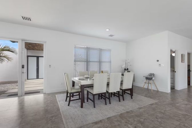 Bright modern dining room with white walls, six-seat rectangular table on an area rug, large window blinds, sliding glass door to a palm-lined patio and a baby high chair.