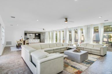 Bright modern open-plan living room with a U-shaped cream leather sectional around a square gray coffee table on a colorful rug, floor-to-ceiling windows with sheer white curtains, tile floors, ceiling fans, and an open kitchen and dining area visible in the background.