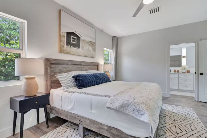 Bright, light-filled modern bedroom with rustic wood platform bed, white linens and navy lumbar pillow, black nightstand with lamp, nature wall art, patterned rug, windows with greenery and doorway to en-suite bathroom.