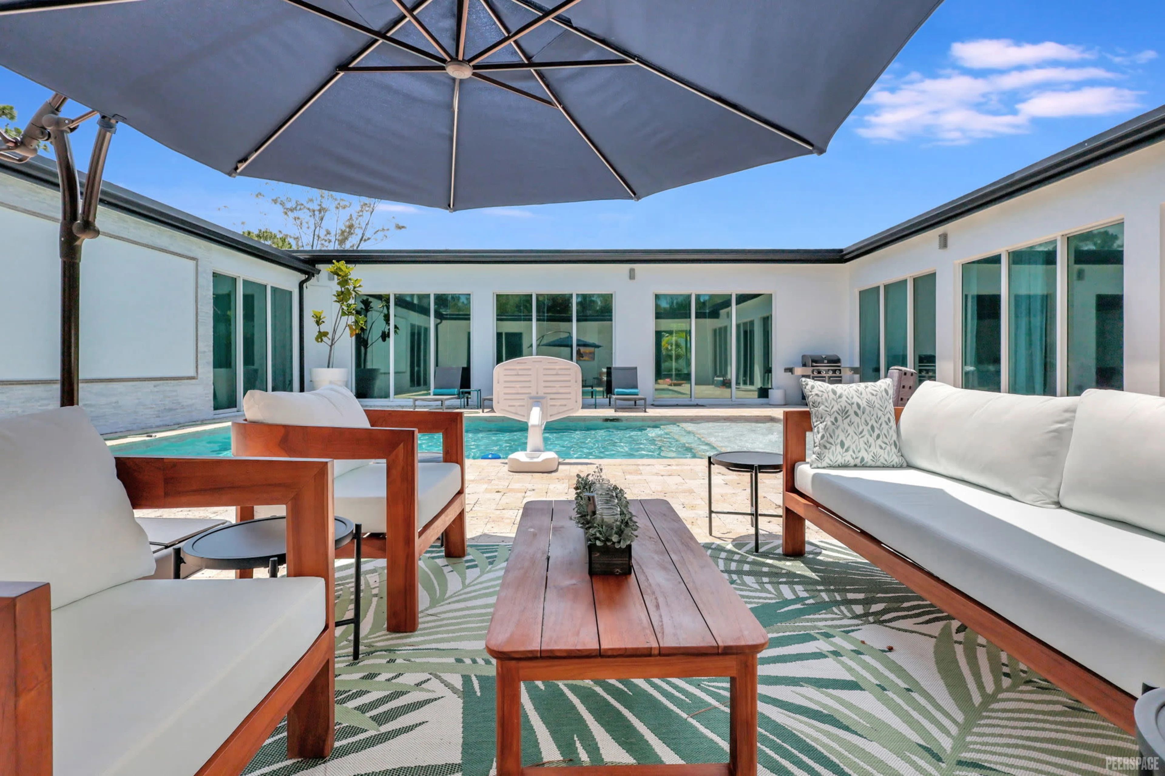 Sunny modern courtyard patio with wooden sofas and chairs, patterned outdoor rug, large navy umbrella and a rectangular swimming pool framed by sliding glass doors