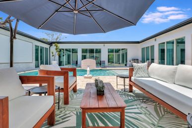 Sunny modern courtyard patio with wooden sofas and chairs, patterned outdoor rug, large navy umbrella and a rectangular swimming pool framed by sliding glass doors