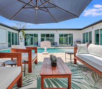 Sunny modern courtyard patio with wooden sofas and chairs, patterned outdoor rug, large navy umbrella and a rectangular swimming pool framed by sliding glass doors