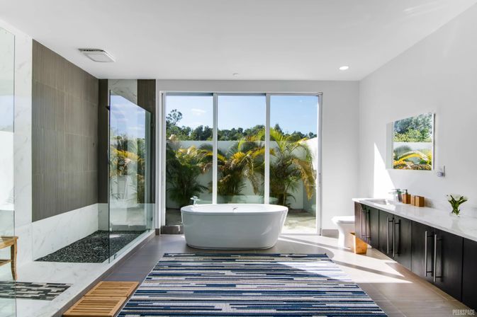 Sunlit modern bathroom with a freestanding white soaking tub centered before floor-to-ceiling sliding glass doors overlooking a palm-lined patio, glass walk-in shower, long dark vanity, and blue-striped rug.