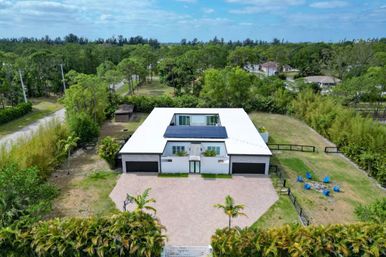 Aerial view of a modern white single-story home with flat roof and solar panels, wide paved driveway and palm trees, fenced grassy yard with blue Adirondack chairs around a fire pit, surrounded by dense greenery in a sunny suburban, tropical setting.