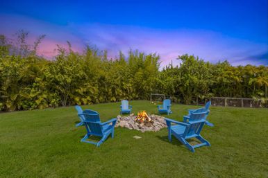 Cozy backyard fire pit at dusk surrounded by six blue Adirondack chairs on a green lawn, framed by lush tropical hedges and a pink-blue sunset sky.