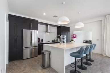 Bright modern open-plan kitchen with a large white island, three blue swivel bar stools, stainless steel fridge and appliances, dark floor-to-ceiling cabinets, two white pendant lights, and a pink orchid on the counter.