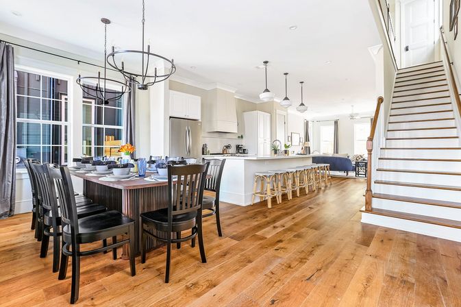 Sunlit open-concept kitchen and dining area with wide-plank hardwood floors, dark wood dining table with black chairs, white island with row of bar stools, pendant lights and staircase