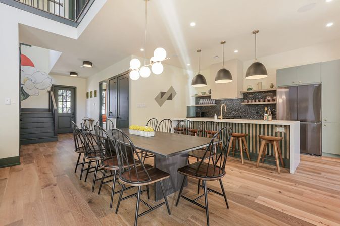 Sunlit open-concept dining room and kitchen with long dark table and eight black spindle-back chairs, globe chandelier, three black pendant lights over a striped island with wooden barstools, stainless fridge, dark speckled backsplash, wide-plank oak floors, staircase with floral mural.