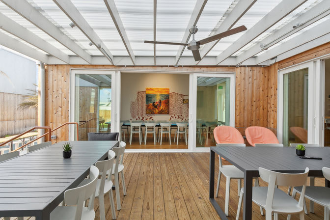 Covered wooden deck patio with modern gray dining tables, white chairs and coral lounge seats, ceiling fan, potted succulents, and sliding glass doors to a dining room with coastal artwork.