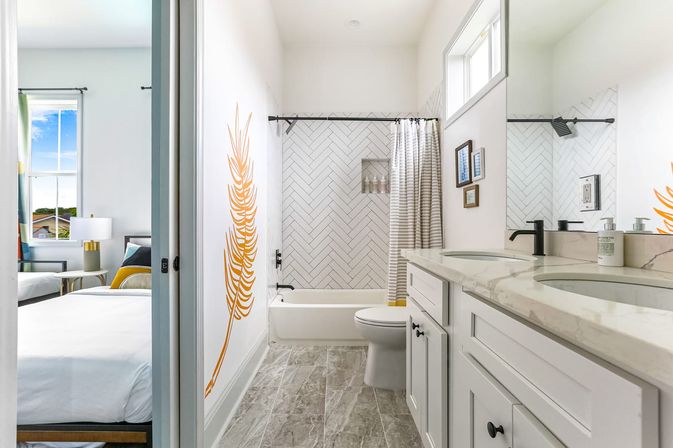 Bright modern residential bathroom with white double vanity and marble countertop, black faucets, bathtub with herringbone subway tile and striped shower curtain, gray floor tile, yellow feather wall decal, and a sunlit bedroom visible through a sliding door.