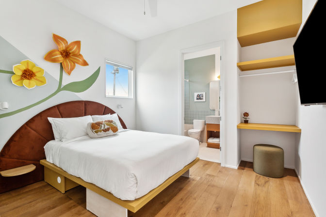 Cheerful modern bedroom with white platform bed, brown semicircular headboard, large yellow-orange flower wall mural, light wood floors, small window, visible en-suite bathroom, built-in yellow shelves and desk, green ottoman and wall-mounted TV.